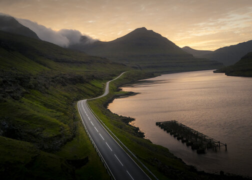 Aerial view of a winding road snakes through verdant hills alongside tranquil waters, reflecting the soft glow of the horizon, Torshavn, Faroe Islands.
