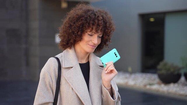 A Cheerful and Smiling Woman with Curly Hair Holding a Green Electronic Device in an Urban Setting