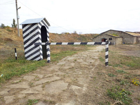 A guard booth painted in black and white stripes. It looks empty and slanted. In front of the booth is a simple wooden barrier, also painted in black and white stripes. It blocks a narrow stone path.