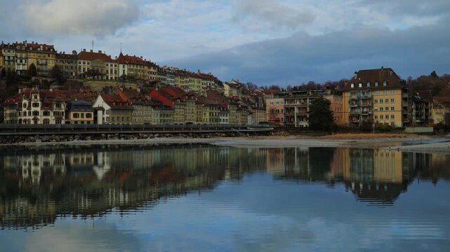 The towering gothic spire of the Bern Minster rises above the historic riverfront houses and the emerald waters of the Aare, captured under a dramatic canopy of gray clouds that emphasize the timeless