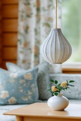 a lampshade in the shape of an ornate indian jali pattern hangs over a table, inside an old cabin with floral printed curtains and pillows on the bed in the background.