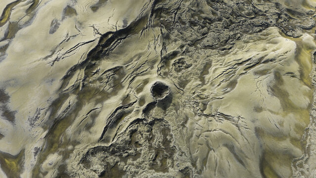 Aerial view of sandy terrain etched with dark lines and a central crater, creating a stark contrast of textures and tones, Selfoss, Iceland.