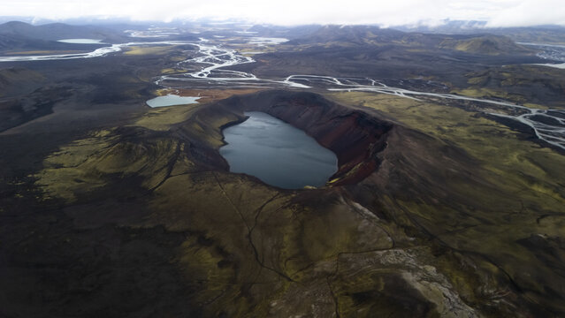 Aerial view of the dramatic contrast of the dark volcanic slopes meeting the serene blue waters of the crater lake, Landmannalaugar, Iceland.