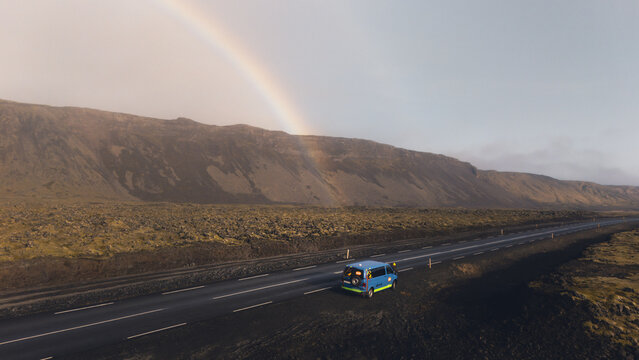 Aerial view of a vibrant blue van driving under a rainbow against the stark Icelandic landscape, the rugged mountains providing a dramatic backdrop, Selfoss, Iceland.