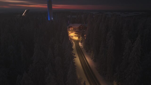 Puijo Tower stands tall in the night sky, illuminated in blue light. The tower is surrounded by tall spruce trees and offers a view of the vast landscape of Kuopio, Finland
