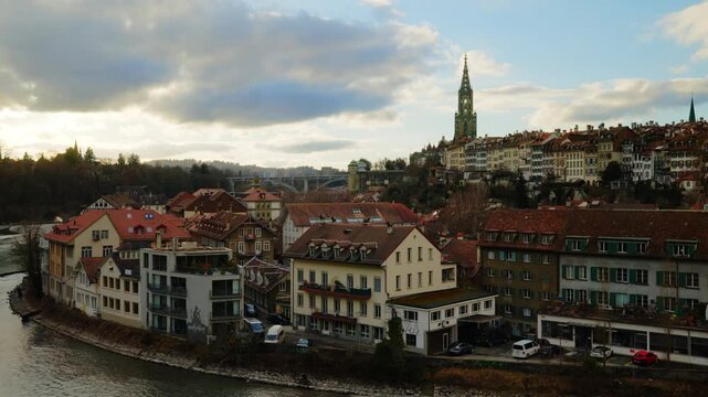The towering gothic spire of the Bern Minster rises above the historic riverfront houses and the emerald waters of the Aare, captured under a dramatic canopy of gray clouds that emphasize the timeless
