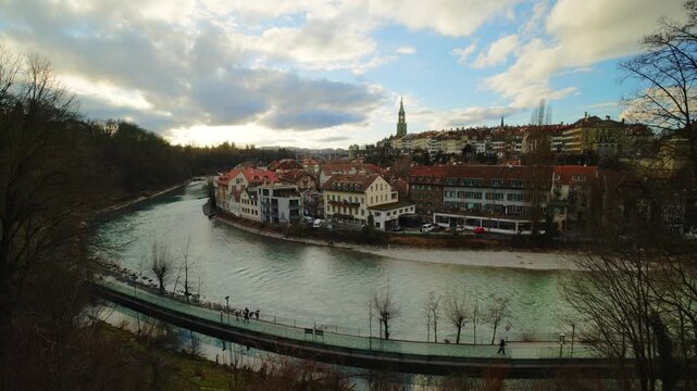 The towering gothic spire of the Bern Minster rises above the historic riverfront houses and the emerald waters of the Aare, captured under a dramatic canopy of gray clouds that emphasize the timeless