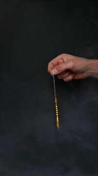Hand using a golden Egyptian Isis pendulum in slow motion with smoke against a black background, vertical video. Symbolic spiritual imagery representing radiesthesia, intuition, subtle energy