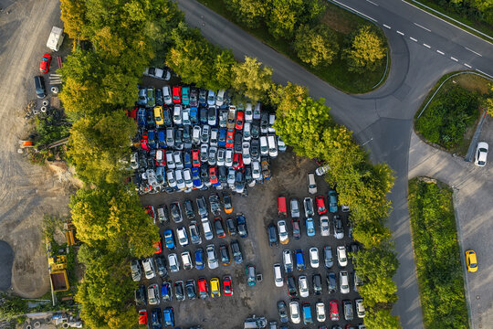 Aerial view of a car graveyard with vehicles parked in rows, bordered by lush green trees and asphalt roads, Mullenbach, Rhineland-Palatinate, Germany.