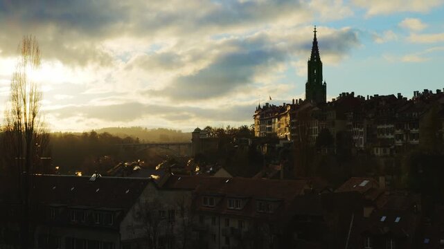 The iconic silhouette of Bern's old town, dominated by the majestic spire of the Minster, is captured in the warm, golden glow of a late afternoon sun, with soft clouds and rising wisps of smoke addin