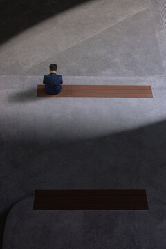 View of a solitary figure sits on a wooden bench, bathed in the interplay of light and shadow, casting dramatic patterns on the concrete floor, Punggol, Singapore.