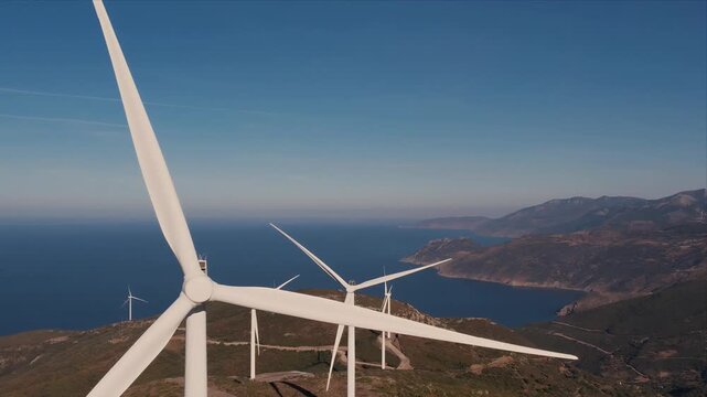 Aerial view of modern wind turbines standing over coastal mountains with the deep blue Aegean Sea beyond. The scene contrasts natural beauty with industrialisation of nature and environmental impact.