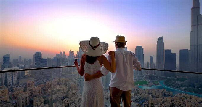 A elegant tourist couple enjoys the romatic sunset behind the skyline of Downtown Dubai, UAE, and cheering with a drink