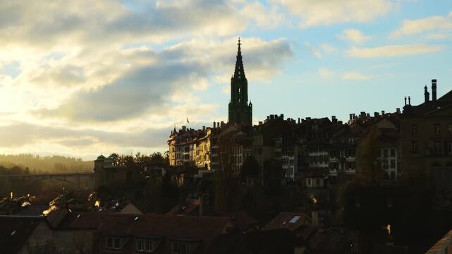 The iconic silhouette of Bern's old town, dominated by the majestic spire of the Minster, is captured in the warm, golden glow of a late afternoon sun, with soft clouds and rising wisps of smoke addin