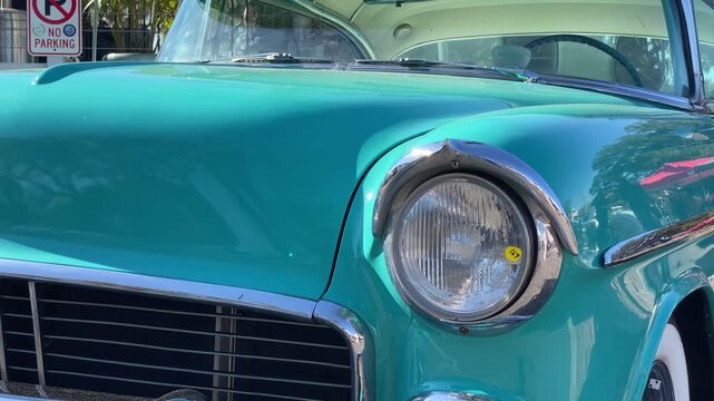 A turquoise classic car is shown in close up with smooth bodywork and chrome trim gleaming in the sun. The vintage headlight and windshield reflect trees and nearby show tents.