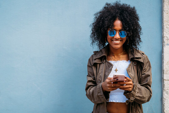 Young woman smiling and texting on smartphone against blue wall