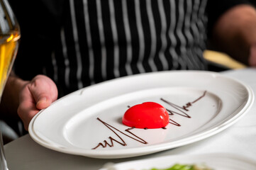 A heart-shaped red glossy mousse dessert with chocolate art is presented on a white oval plate by a chef or waiter in a restaurant setting
