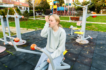 Teenage girl resting on outdoor fitness equipment, hand on face, feeling tired after workout, recovery moment, healthy lifestyle and wellbeing concept
