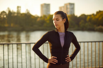 Fototapeta premium Woman runner resting by river after training. Hands on hips, the female athlete pauses on a riverside path at dusk, cooling down and showing confidence. Recovery and focus sport fitness concept.