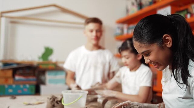 Smart teenager modeling cup of clay while laughing at art lesson. Diverse happy student standing while doing creative activity at workshop. Creative activity concept. Blurred background. Edification