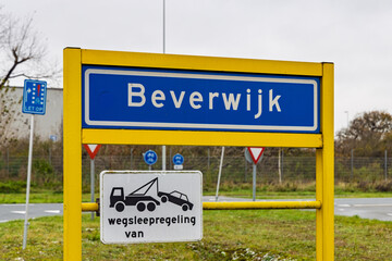 A blue city entrance sign for Beverwijk, Netherlands, featuring a yellow frame and a towing zone warning sign against a blurred background.