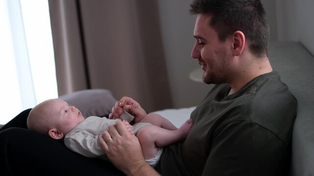 The middle plan of the portrait is in warm colors of a young father and newborn son. A father plays and kisses his little son on the bed. The concept of love, happy fatherhood.