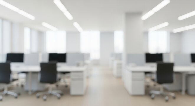 A blurred view of a bright modern open-plan office with multiple empty white desks black ergonomic chairs and dark computer monitors under bright ceiling lights