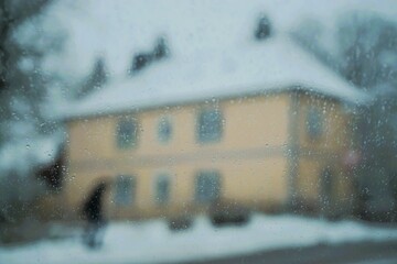 Abstraktes Motiv mit Schneelandschaftspanorama mit Hauptstra&szlig;e, Person auf Gehweg und altem beigen Haus vor hohen Tannen und Himmel bei Schneefall, Eis, Sturm und Frost im Winter