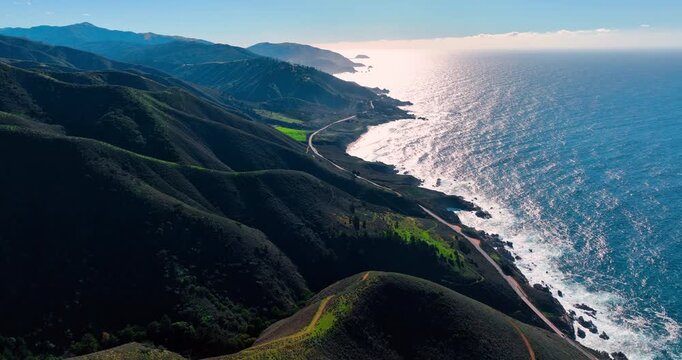 Flight over spectacular ridges mountains with a highway along the foot. Sun dazzles the waterscape of the Pacific Ocean. Big Sur, California, USA.