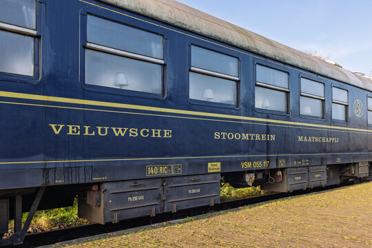 Side view of a blue vintage passenger wagon of the Veluwsche Stoomtrein Maatschappij, showing gold lettering and windows with interior lamps. Beekbergen Netherlands, 7 November 2025.