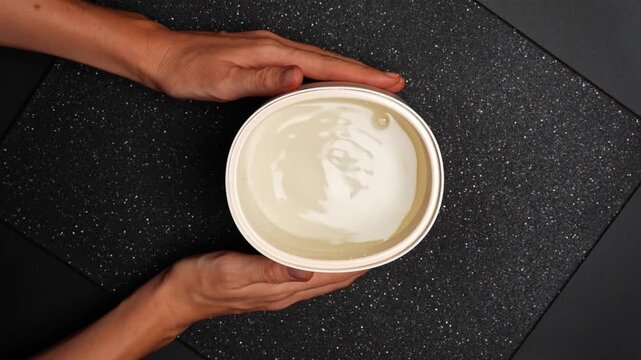 Person holds a round container filled with liquid on a dark surface. A person is holding a round container filled with a creamy liquid on a flat dark surface.