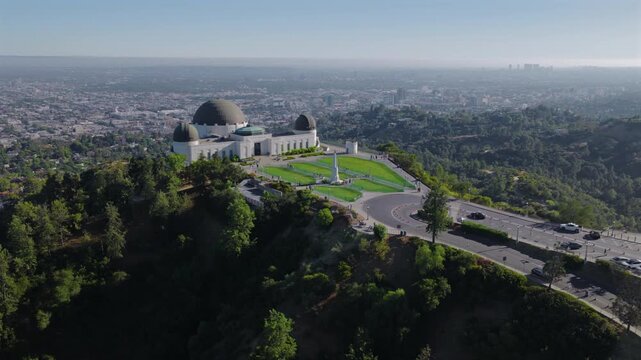 Aerial view of Griffith Observatory above Los Angeles California with city skyline, green hills, roads and traffic. Iconic landmark captured by drone in clear daylight conditions.