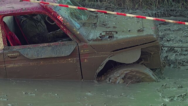 An extreme off-road competition. A red utility vehicle, covered in brown mud is skidding, but is being moved by a winch through a deep muddy puddle. The driver inside, navigating the challenge