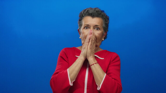 Senior woman with grey hair in a red sweater looks shocked against a vibrant blue background, expressing surprise and emotion vividly captured in this stock image.