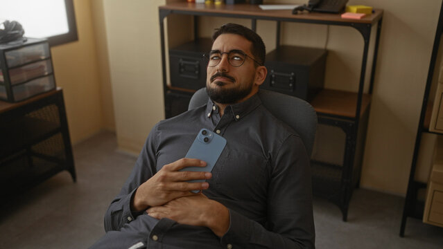 Young man with beard relaxing in an office chair while using smartphone indoors, surrounded by shelves and storage boxes, showcasing a modern workplace environment.