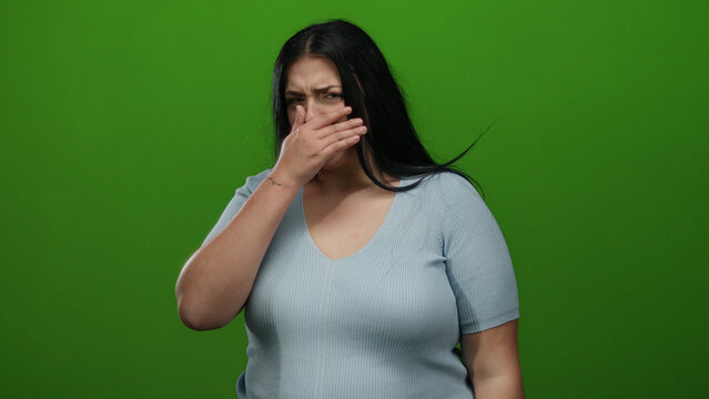 Young woman covering mouth on green background, expressing surprise or embarrassment, dressed in blue shirt, isolated and looking concerned, suitable for emotional themes.