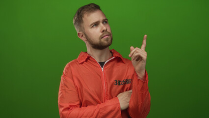 Fototapeta premium Young caucasian prisoner man in numbered orange jumpsuit pointing right index finger upward in studio with green backdrop while arm is folded across chest; contemplation.