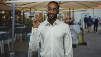 Obraz premium Man points finger upward on street cafe terrace near restaurant while wearing glasses and white shirt; contemplative reflection.