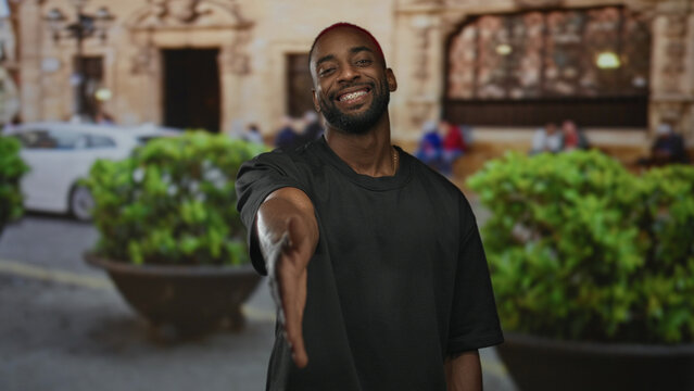 Man offers outstretched hand for handshake on a city street in front of building, smiling and leaning forward; friendly greeting.