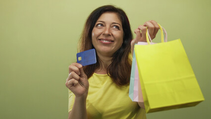 Middle-aged hispanic woman holds creditcard and pastel shopping bag in green studio setting; shopping joy.