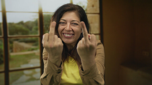 Woman in yellow shirt shows two middle finger gestures in room with window grid and sunlight streaming through; defiance.