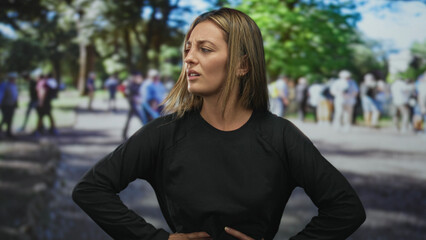 Fototapeta premium Young blonde woman standing with hands on hips squinting and pressing hand to stomach on street amid blurred crowd; frustration resolve.