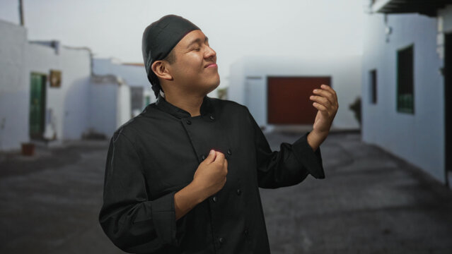 Hispanic man chef mimes air guitar with hands on a sunlit street between white buildings; playful celebration.