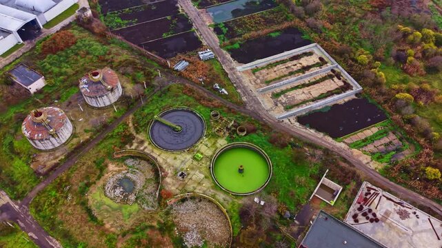 Aerial view reveals an abandoned industrial site featuring circular tanks surrounded by greenery and patches of land. The area shows signs of neglect and decay.