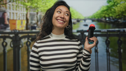 Young woman holding a game controller and smirking on a canal street bridge in amsterdam, outdoors  joy playful travel. © Krakenimages.com