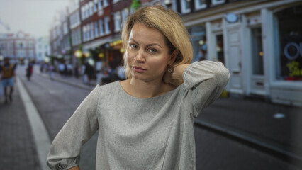 Fototapeta premium Young blonde woman lightly touches her neck outside a brown brick building on a busy street; contemplation.