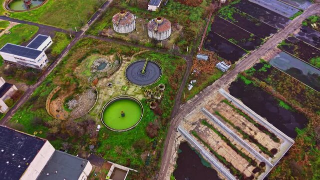 Aerial view shows abandoned tanks surrounded by green water and empty structures. The site features scattered greenery and layout of old infrastructure under a clear sky.