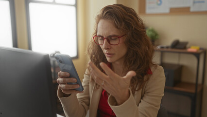 Woman in office using smartphone with confused expression, wearing red glasses, seated at a desk with a modern interior, suggesting business communication or technology use.