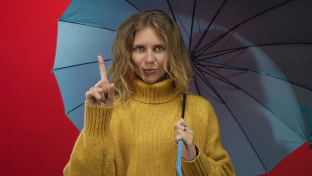 In this creative portrait, woman hand counting props appears against studio walls next to a blonde holding an umbrella while smiling and displaying a finger gesture that feels happy with two symbols.