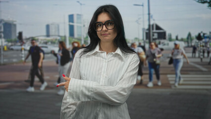 Fototapeta premium Young hispanic woman wearing glasses points finger to crosswalk on urban street among pedestrians; choice uncertainty doubt.
